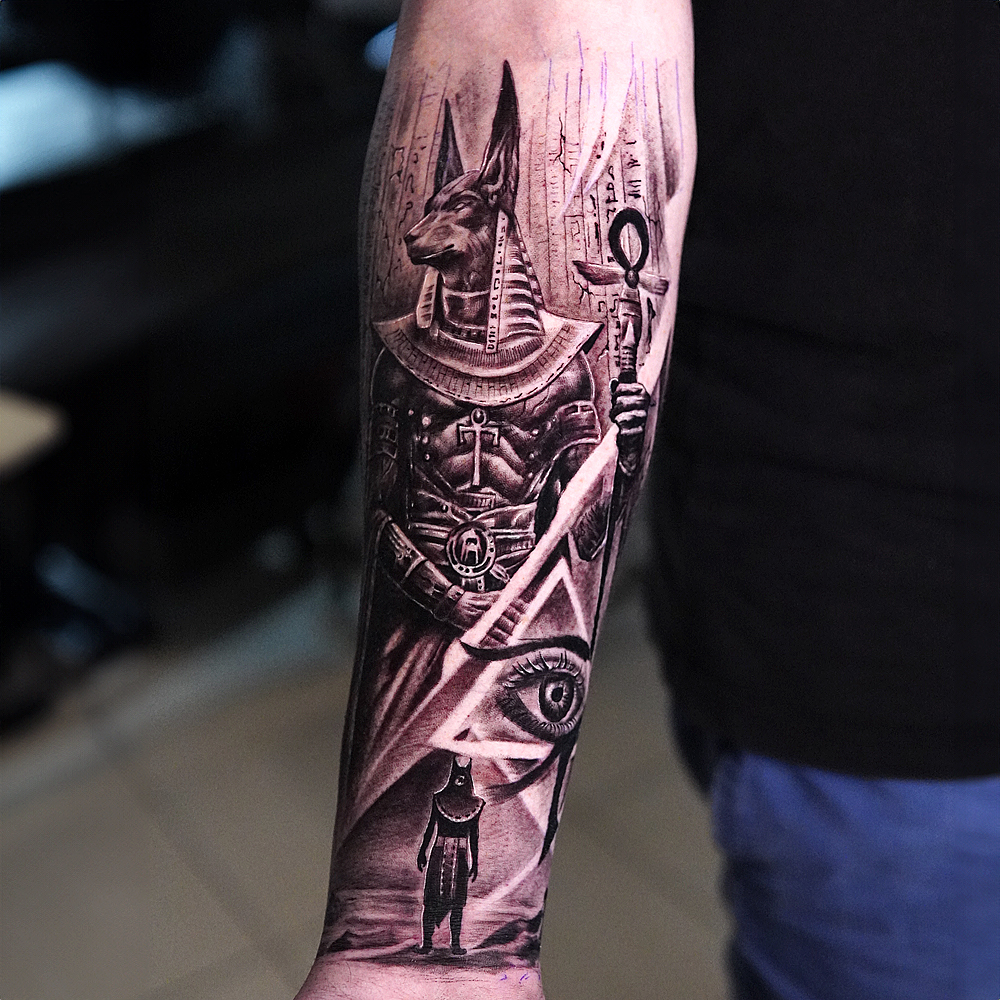 Anubis Forearm Tattoo Design Black and grey forearm tattoo featuring Anubis, an ankh symbol, and an eye inside a triangle with Egyptian hieroglyphics.